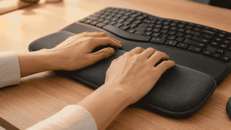 Hands resting lightly on a cushioned wrist rest in front of an ergonomic keyboard during a typing pause