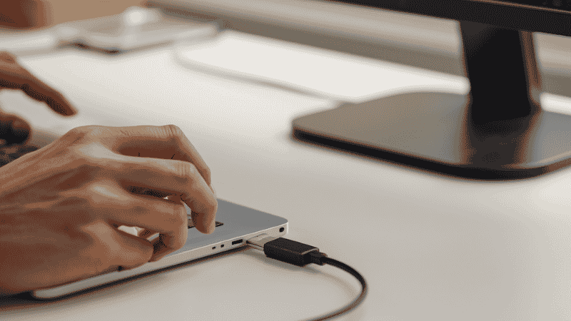 A hand plugging a single Thunderbolt cable into a laptop on a home office desk with all peripherals already connected through the dock