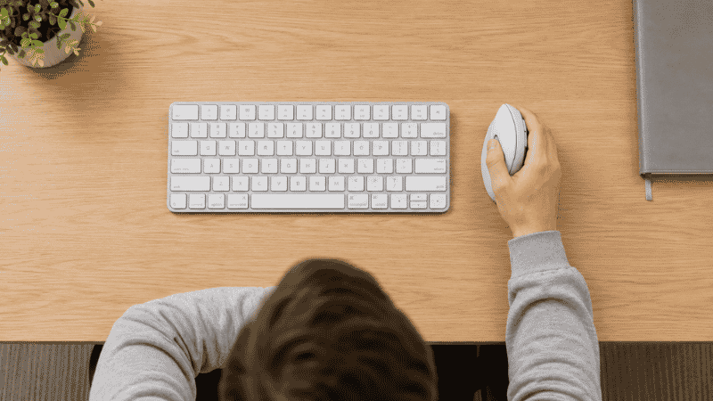 Top-down view of a home office desk showing a vertical mouse positioned directly beside the keyboard with elbow at 90 degrees