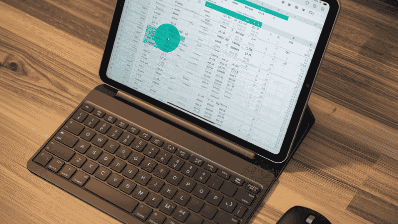 A modern iPad Pro desk setup, featuring a mechanical keyboard and a Logitech MX mouse, with the screen displaying the iPadOS circular cursor navigating a spreadsheet.