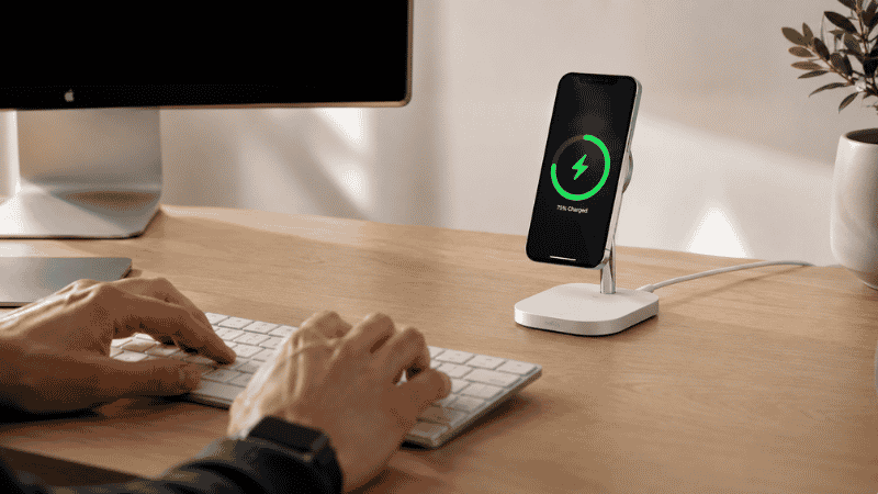 A wireless charging stand on a clean home office desk with an iPhone charging magnetically while the owner types on a keyboard